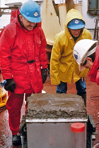 John and others in raingear inspect a box corer sample on the deck of a ship